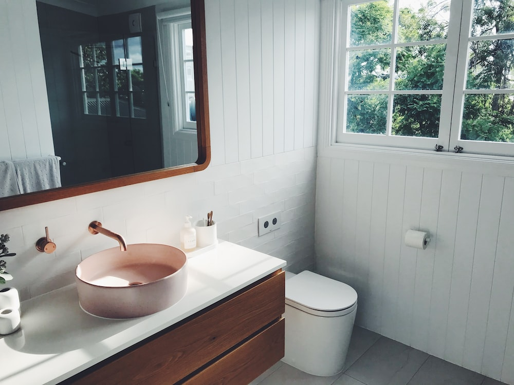 a vintage bathroom with a soft pink sink, wooden mirror, and glass windows